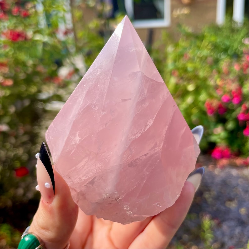 May include: A large, pale pink rose quartz crystal in the shape of a pyramid. The crystal is smooth and polished, with a slight translucence. It is being held in a hand.
