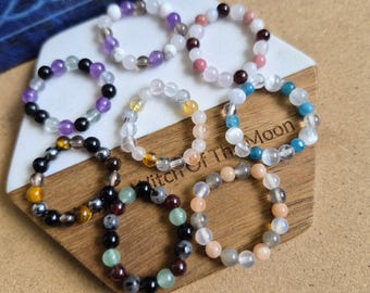 May include: Eight circular rings made of small, polished gemstone beads in various colors including purple, white, black, pink, blue, yellow, and green. The rings are arranged on a hexagonal display board with the text Witch of the Moon engraved.