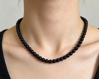 May include: A single-strand necklace composed of uniform, polished black spherical beads. The necklace sits closely around the neck, featuring a consistent arrangement of smooth, dark, circular beads that reflect light across their glossy surfaces.