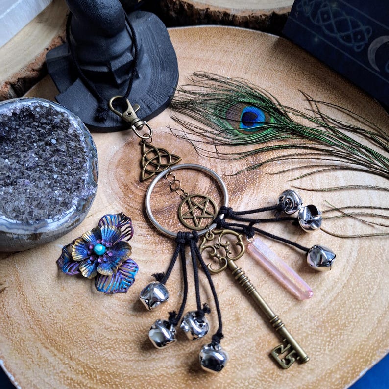 Puede incluir: Un amuleto de llave plateado con un pentagrama, un nudo celta y un colgante de cristal. El amuleto est&aacute; unido a un cord&oacute;n negro con cascabeles plateados y una pluma de pavo real.