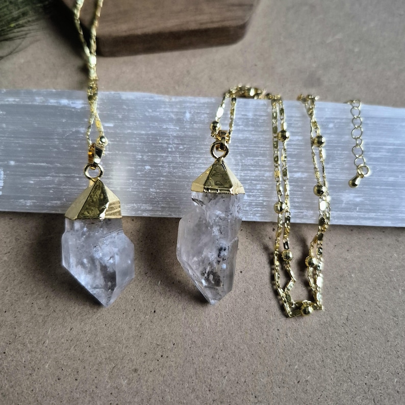 May include: Two necklaces featuring raw, clear quartz crystal points capped in polished gold-tone metal. Each pendant hangs from a delicate gold-tone chain with small, evenly spaced metallic beads, finished with a short adjustable extender chain.