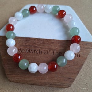 May include: A colourful beaded bracelet with red, pink, green, and white gemstones. The round beads are strung together. The bracelet is displayed on a wooden stand with the text "White Witch Of The Moon".