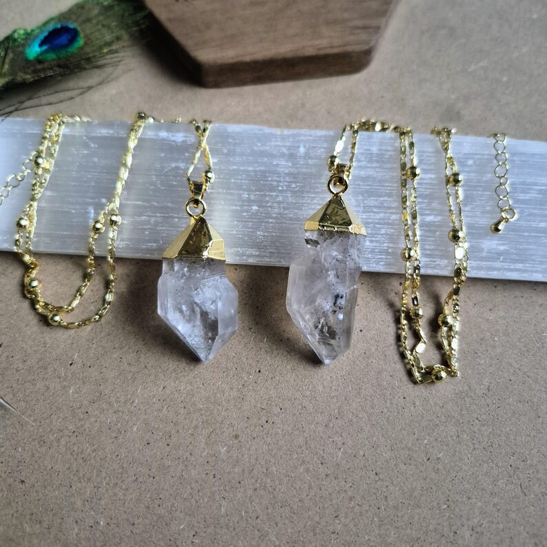 May include: Two necklaces featuring raw, clear quartz crystal points capped in gold-toned metal. Each pendant hangs from a delicate gold-toned chain with small, evenly spaced beads. The crystals are translucent with natural internal inclusions.