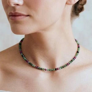 May include: A beaded necklace featuring small, round, faceted stones in alternating shades of deep magenta, forest green, and black. The necklace sits closely around the neck, showcasing a uniform pattern of polished, multi-coloured gemstone beads.