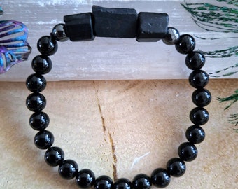 May include: A black onyx beaded bracelet with a black obsidian focal bead. The bracelet is on a white surface.