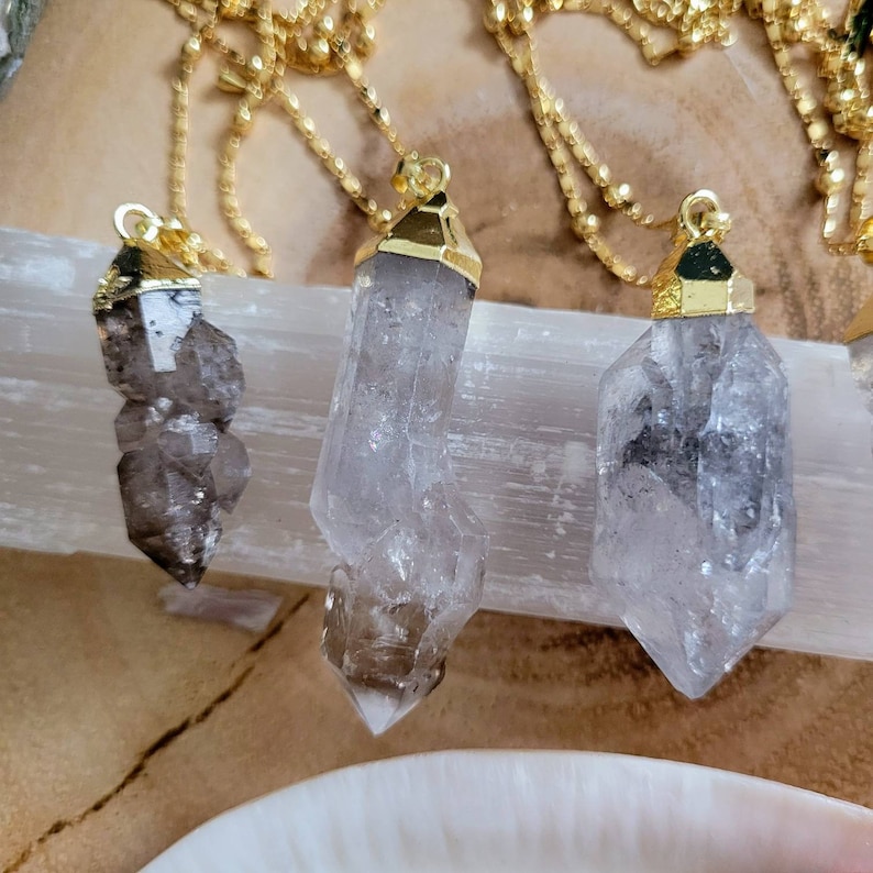 May include: Three crystal point pendants with gold-toned metal caps, each suspended from a delicate gold-toned chain. The crystals are translucent with dark inclusions and natural facets, showcasing varied shapes and sizes in a vertical arrangement.