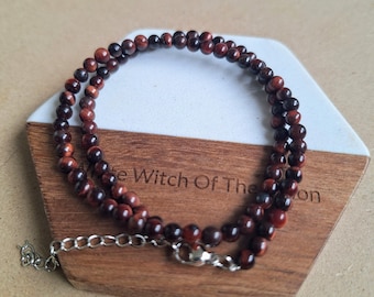 May include: A layered necklace made of small, round, reddish-brown beads. The necklace is displayed on a wooden and white hexagonal coaster. The necklace has a silver-colored clasp and chain.