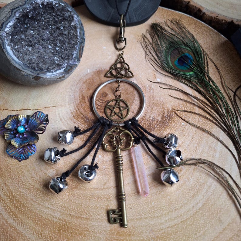 Puede incluir: Un amuleto de llave de plata con un pentagrama y un triquetra, colgando de un anillo de plata con borlas negras y cascabeles plateados. El amuleto est&aacute; sobre una superficie de madera con una pluma de pavo real.