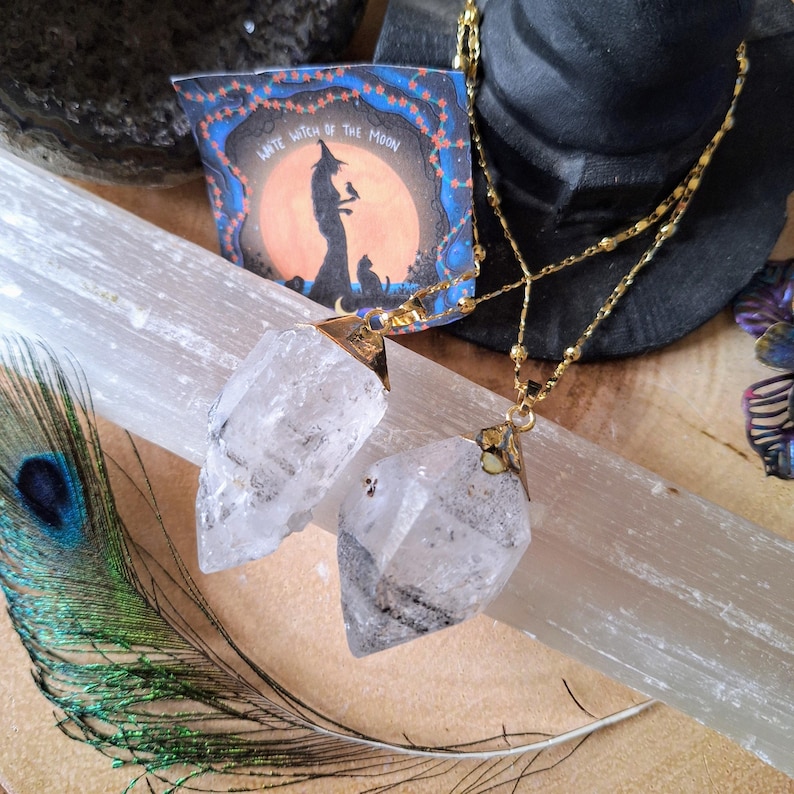 May include: Two clear, raw crystal pendants hang from gold-toned chains. Each crystal has a pointed, faceted shape with natural inclusions. A small rectangular card is visible behind the pendants, featuring an illustration of a witch and the text "Witch of the Moon" printed in white against an orange and blue background.