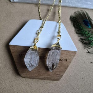 May include: Two clear, raw crystal pendants suspended from delicate gold-toned chains. Each crystal is capped with a gold-toned metal setting at the top, featuring a faceted, pointed shape that hangs vertically from the thin, metallic link chains.