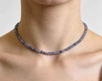 May include: A choker necklace featuring a single strand of small, faceted, translucent blue-grey beads. The beads are uniform in size and shape, creating a continuous circular design that rests closely around the base of the neck.