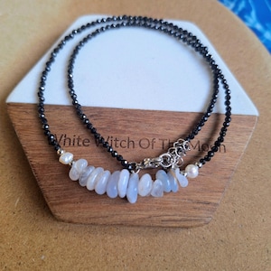 May include: A necklace with a strand of small, black beads. The necklace has a central row of light blue stones and small white pearls. The necklace is displayed on a wooden hexagonal stand.