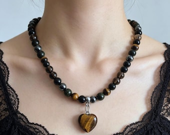 May include: A beaded necklace featuring alternating round black, dark brown, and metallic gray stones. The necklace includes a central heart-shaped pendant made of brown and gold tiger eye stone, suspended from a silver-toned metal jump ring.