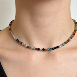 May include: A choker necklace featuring a single strand of small, faceted, multi-coloured gemstone beads. The beads display a variety of hues including red, orange, yellow, green, blue, and black, arranged in a repeating pattern around the neck.