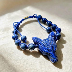 May include: A bracelet featuring a carved blue stone wolf head focal piece and round blue stone beads strung on a blue cord. The design includes an adjustable sliding knot closure with a small gold-toned metal accent bead on the cord.