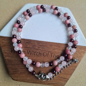 May include: A layered necklace featuring small, round beads in shades of pink, red, and clear. The necklace is displayed on a wooden hexagonal surface. The text "White Witch Of The" is partially visible.