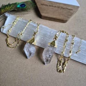 May include: Two necklaces featuring clear, pointed crystal pendants capped in gold-toned metal. Each pendant hangs from a delicate gold-toned chain with small, evenly spaced beads. The necklaces are displayed on a long, rectangular piece of white selenite.