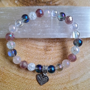 May include: A bracelet made of iridescent blue, clear, and pink beads with a silver heart charm with wings. The bracelet is on a white surface.