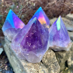 May include: Four amethyst crystal points with iridescent coatings. The crystals are arranged in a pyramid shape on a rock surface.