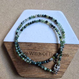 May include: A delicate necklace with small, faceted green beads. The necklace is double-stranded and has a silver clasp. It is displayed on a wooden and white hexagonal stand. The text "White Witch Of The Moon" is visible.