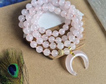 May include: A beaded necklace featuring smooth, pale pink rose quartz spheres strung together. The necklace includes a gold-toned spacer bead with small clear crystals and a large, crescent-shaped rose quartz pendant attached with a gold-toned metal bail.