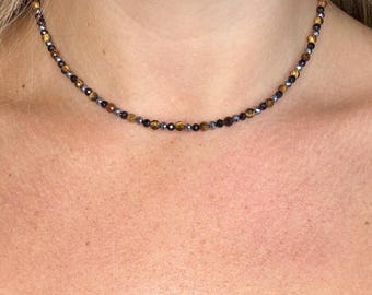 May include: A beaded necklace with small, round beads in shades of brown, gold, and gray. The necklace is worn around the neck, showcasing the delicate design and color variations. The beads appear to be made of natural stone.