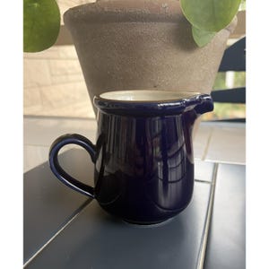 May include: A small, deep navy blue ceramic creamer pitcher with a glossy finish, a curved handle on the side, and a small pouring spout. The interior of the pitcher is a contrasting off-white color, and the base is rounded with a smooth, solid design.
