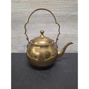 May include: A polished brass teapot with a rounded body, a matching lid featuring a small knob handle, and a curved spout. The teapot includes a tall, decorative arched handle that is hinged at the sides, showcasing a metallic gold finish.
