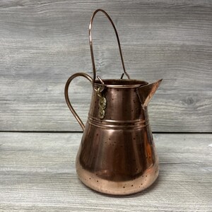 May include: A copper-colored pitcher with a curved handle and a spout. The pitcher has a small handle on the side and a larger handle that arches over the top. The pitcher is made of copper and has a shiny finish. The pitcher is likely used for pouring liquids.