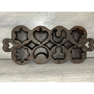 May include: A cast iron muffin pan with various shapes, including a star, heart, rooster, and clover. The pan has a dark, rustic finish and handles on each end. The pan is designed for baking decorative muffins.