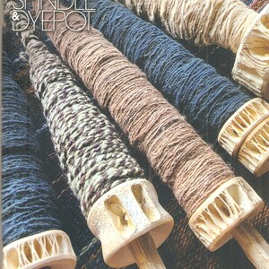 Può includere: Primo piano di diversi fusi avvolti con filato di vari colori, tra cui beige, marrone e blu. I fusi hanno cime decorative in legno. L'immagine è tratta dalla rivista "Shuttle Spindle & Dyepot".