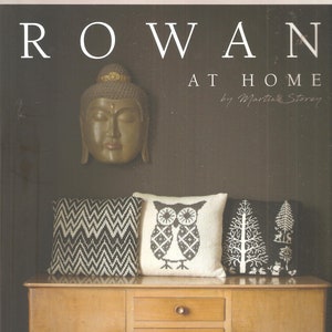 May include: A brown wooden dresser with three drawers and two cabinet doors. The dresser has a light wood finish and brass knobs. Three decorative pillows are on top of the dresser. The pillows are white with black designs. The pillows feature a geometric pattern, an owl, and a tree design. The text "ROWAN AT HOME by Martin Storey" is on the wall behind the dresser.