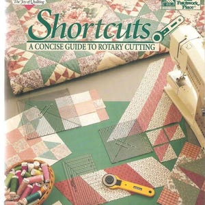 May include: A book titled "Shortcuts: A Concise Guide to Rotary Cutting" by Donna Lynn Thomas. The cover features a quilt with floral and geometric patterns. Sewing tools, fabric pieces, and spools of thread are arranged on a green surface, suggesting a quilting theme.