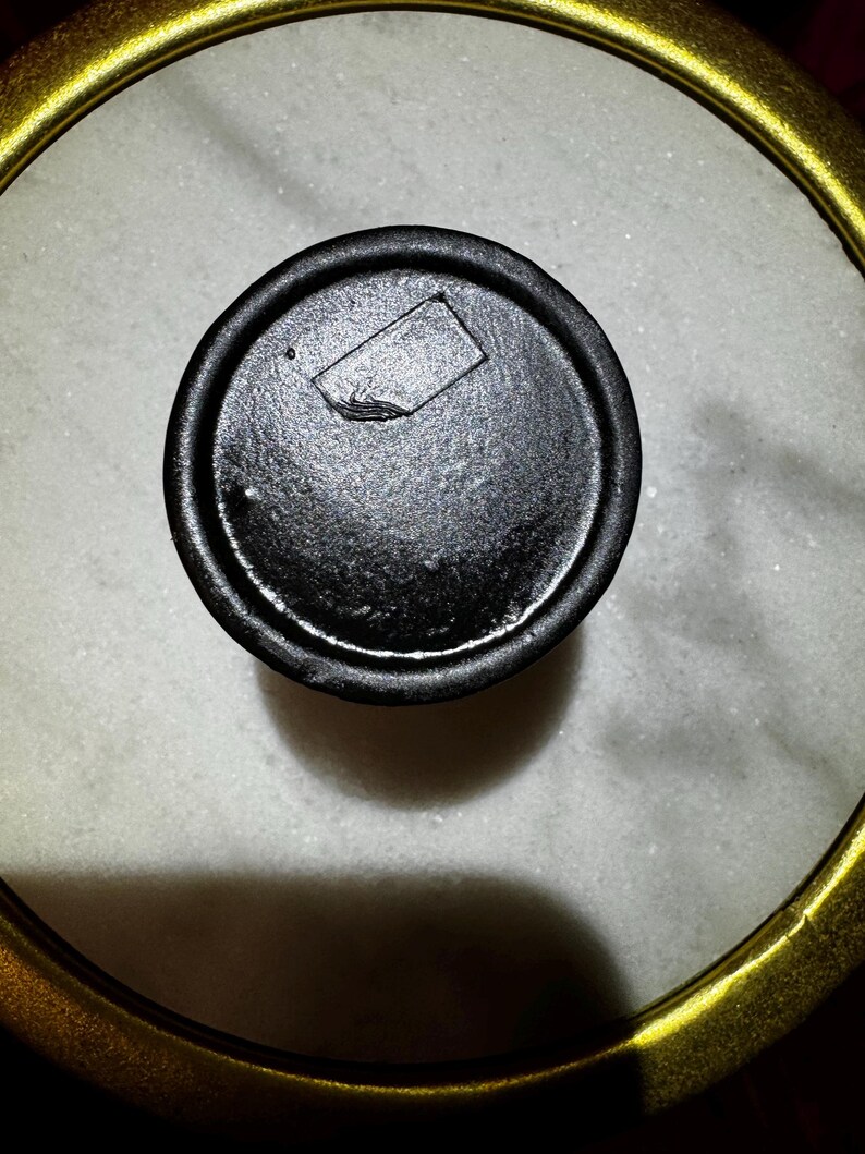 K&ouml;nnte beinhalten: Nahaufnahme eines schwarzen Deckels mit rechteckiger Vertiefung, vor einem wei&szlig;en, gesprenkelten Hintergrund. Der Deckel ist von einem goldfarbenen, kreisf&ouml;rmigen Rahmen umgeben. Das Bild ist gut beleuchtet und hebt die Texturen und Formen hervor.
