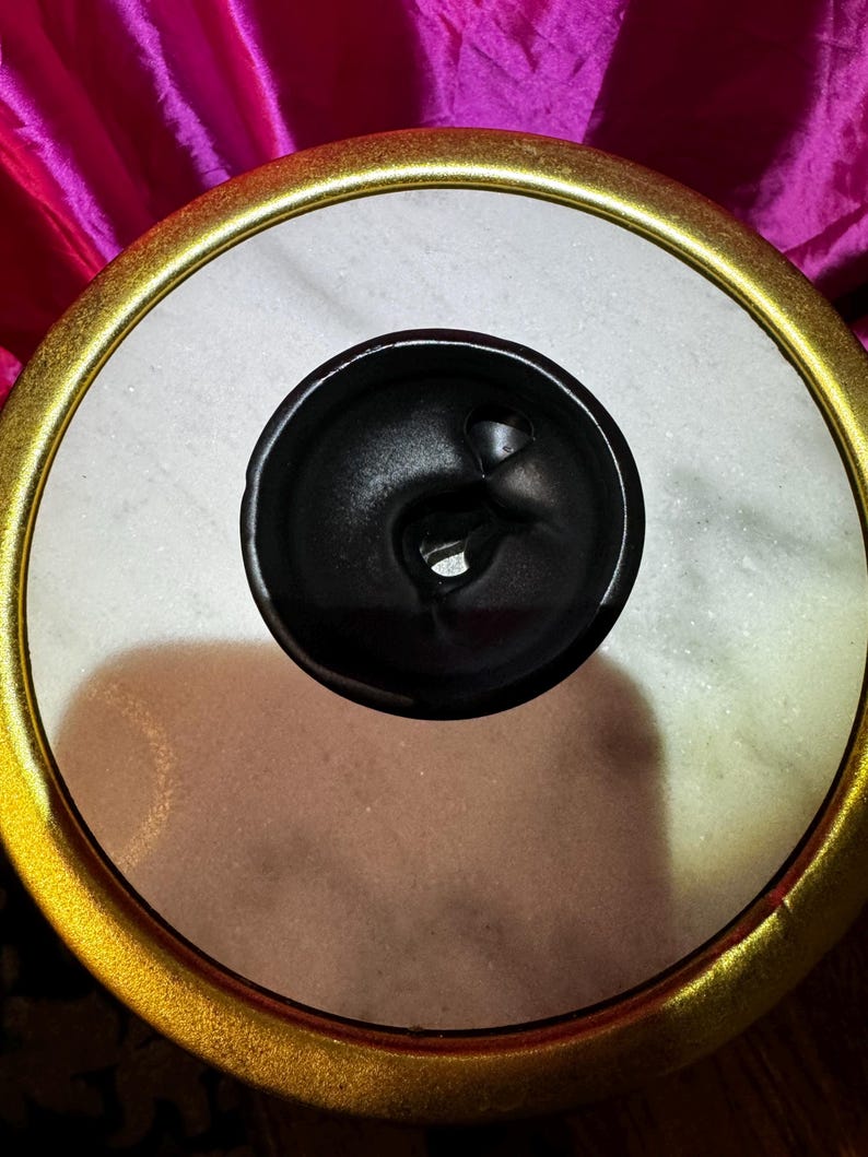 K&ouml;nnte beinhalten: Ein dekoratives, rundes Objekt mit einem goldfarbenen Rand und einem schwarzen Zentrum. Das Zentrum hat ein einzigartiges, wirbelndes Design. Das Objekt befindet sich vor einem Hintergrund aus leuchtend pinkfarbenem Stoff.