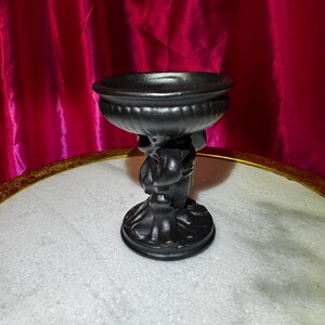 K&ouml;nnte beinhalten: Eine schwarze dekorative Schale, die von einer Cherubfigur getragen wird. Die Schale hat ein breites, flaches Design mit einem abgerundeten Rand. Der Cherub h&auml;lt die Schale &uuml;ber seinem Kopf. Der Sockel ist rund und detailliert. Der Hintergrund ist ein roter Vorhang.