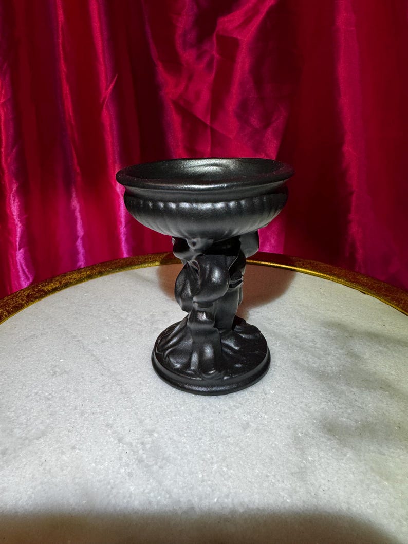 K&ouml;nnte beinhalten: Eine schwarze dekorative Schale, die von einer Cherubfigur getragen wird. Die Schale hat ein breites, flaches Design mit einem abgerundeten Rand. Der Cherub h&auml;lt die Schale &uuml;ber seinem Kopf. Der Sockel ist rund und detailliert. Der Hintergrund ist ein roter Vorhang.