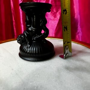 K&ouml;nnte beinhalten: Ein schwarzer dekorativer Kerzenhalter mit einem detaillierten Design mit Figuren. Der Kerzenhalter ist etwa 7,6 cm hoch. Der Hintergrund umfasst einen rosa Stoff und eine wei&szlig;e Oberfl&auml;che.