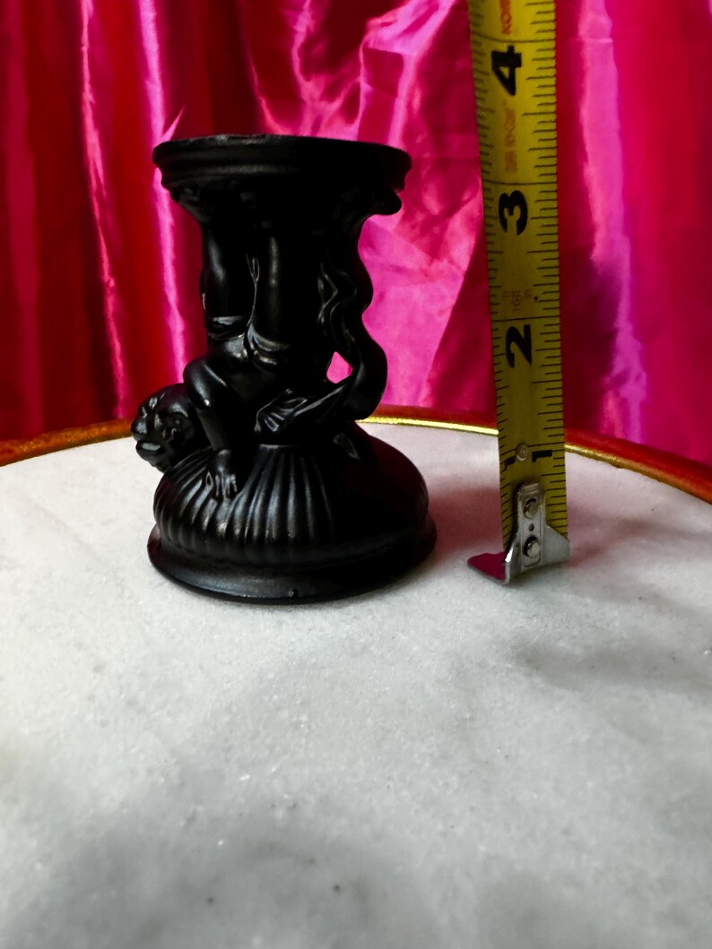 K&ouml;nnte beinhalten: Ein schwarzer dekorativer Kerzenhalter mit einem detaillierten Design mit Figuren. Der Kerzenhalter ist etwa 7,6 cm hoch. Der Hintergrund umfasst einen rosa Stoff und eine wei&szlig;e Oberfl&auml;che.