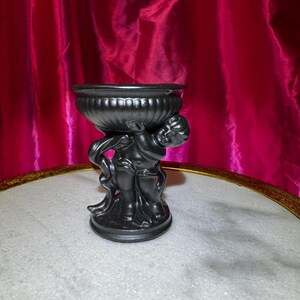 K&ouml;nnte beinhalten: Eine schwarze dekorative Urne mit Cherubim-Sockel. Die Urne hat eine breite, geriffelte Schale und wird von zwei Cherubim-Figuren getragen. Die Cherubim halten die Schale hoch. Die Urne besteht aus dunklem Material und hat eine matte Oberfl&auml;che.