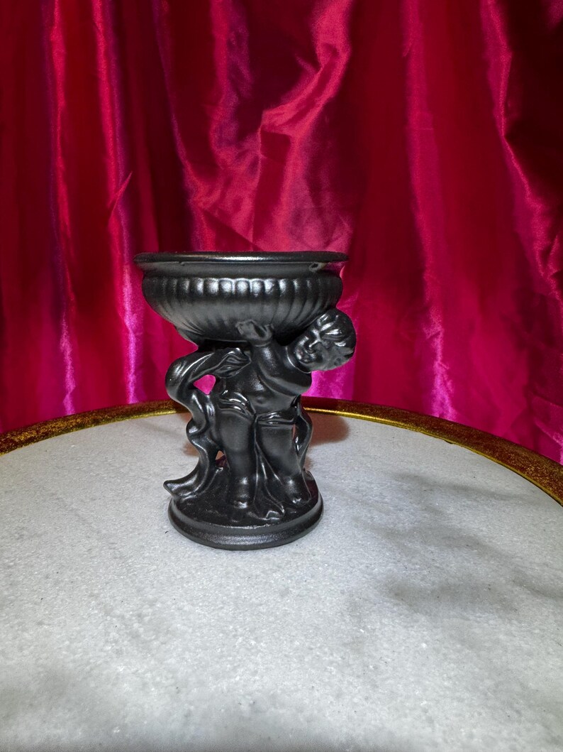 K&ouml;nnte beinhalten: Eine schwarze dekorative Urne mit Cherubim-Sockel. Die Urne hat eine breite, geriffelte Schale und wird von zwei Cherubim-Figuren getragen. Die Cherubim halten die Schale hoch. Die Urne besteht aus dunklem Material und hat eine matte Oberfl&auml;che.