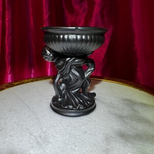 K&ouml;nnte beinhalten: Eine schwarze dekorative Urne mit geriffeltem schalenf&ouml;rmigem Oberteil, getragen von einer skulptierten Figur. Die Urne hat eine dunkle, metallische Oberfl&auml;che und steht auf einem runden Sockel. Der Hintergrund zeigt einen roten Vorhang.