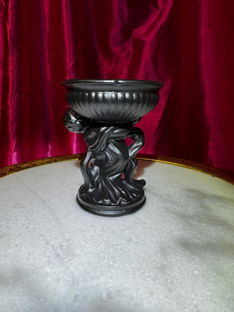 K&ouml;nnte beinhalten: Eine schwarze dekorative Urne mit geriffeltem schalenf&ouml;rmigem Oberteil, getragen von einer skulptierten Figur. Die Urne hat eine dunkle, metallische Oberfl&auml;che und steht auf einem runden Sockel. Der Hintergrund zeigt einen roten Vorhang.