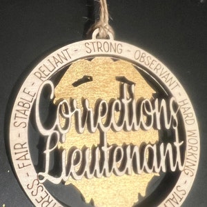 May include: A wooden ornament with a gold background and the words "Corrections Lieutenant" in a cutout design. The ornament is surrounded by a circle of text that reads "Stalwart - Calm Under Stress - Fair Stable Reliant - Strong - Observant - Hard Working".