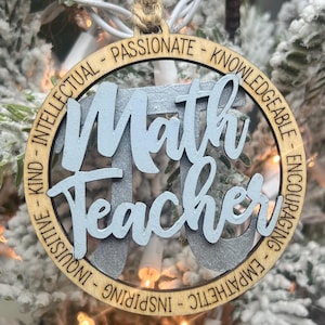 May include: A wooden Christmas ornament with the words "Math Teacher" written in blue paint. The ornament is decorated with a circle of words describing positive qualities, including "intellectual", "passionate", "knowledgeable", "inspiring", "empathic", "inquisitive", "kind", and "encouraging".
