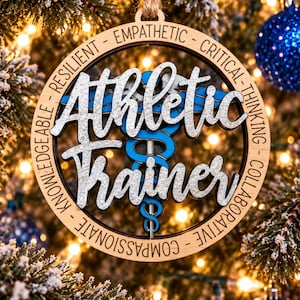 May include: A circular wooden ornament featuring the text Athletic Trainer in silver glitter script. A blue medical caduceus symbol is centered behind the text. The outer wooden ring is engraved with the words Empathetic, Critical, Thinking, Collaborative, Compassionate, Knowledgeable, and Resilient.