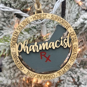 May include: A wooden Christmas ornament with a gray mortar and pestle design. The ornament is inscribed with the word "pharmacist" and the text "Problem Solving - Honest - Supportive - Detail Oriented - Helpful - Communicative - Observant".