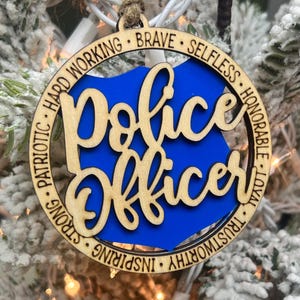 May include: Wooden Christmas ornament with a blue background and the words "Police Officer" in a cursive font. The ornament is surrounded by a circle with the words "Trustworthy, Loyal, Honorable, Selfless, Brave, Hard Working, Inspiring, Strong, Patriotic" written around it.