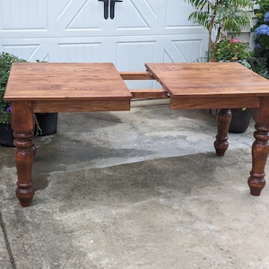 May include: A brown wooden table with a leaf extension. The table has turned legs with a decorative design. The table is sitting on a concrete patio.