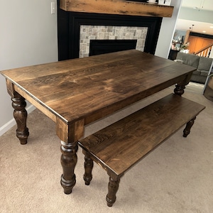 May include: A dark stained wooden dining table with a matching bench. The table has a rectangular top and turned legs. The bench is smaller, with a similar design. The furniture is set on a light-colored carpet.